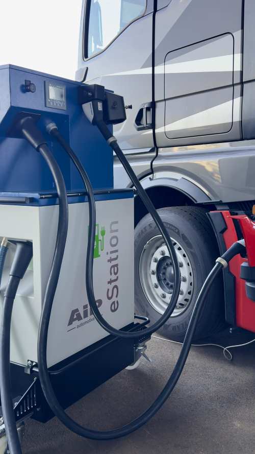 Ladestation mit mehreren Kabeln vor einem silbernen Lkw mit schwarzem Reifen und rotem Unterbau.