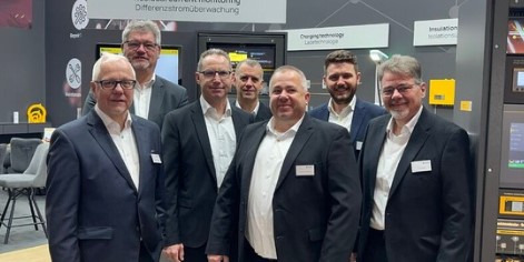 Sieben Personen in Anzügen stehen in einer Gruppe. Alle tragen Namensschilder. Im Hintergrund sind technische Geräte und Informationsschilder zu sehen.